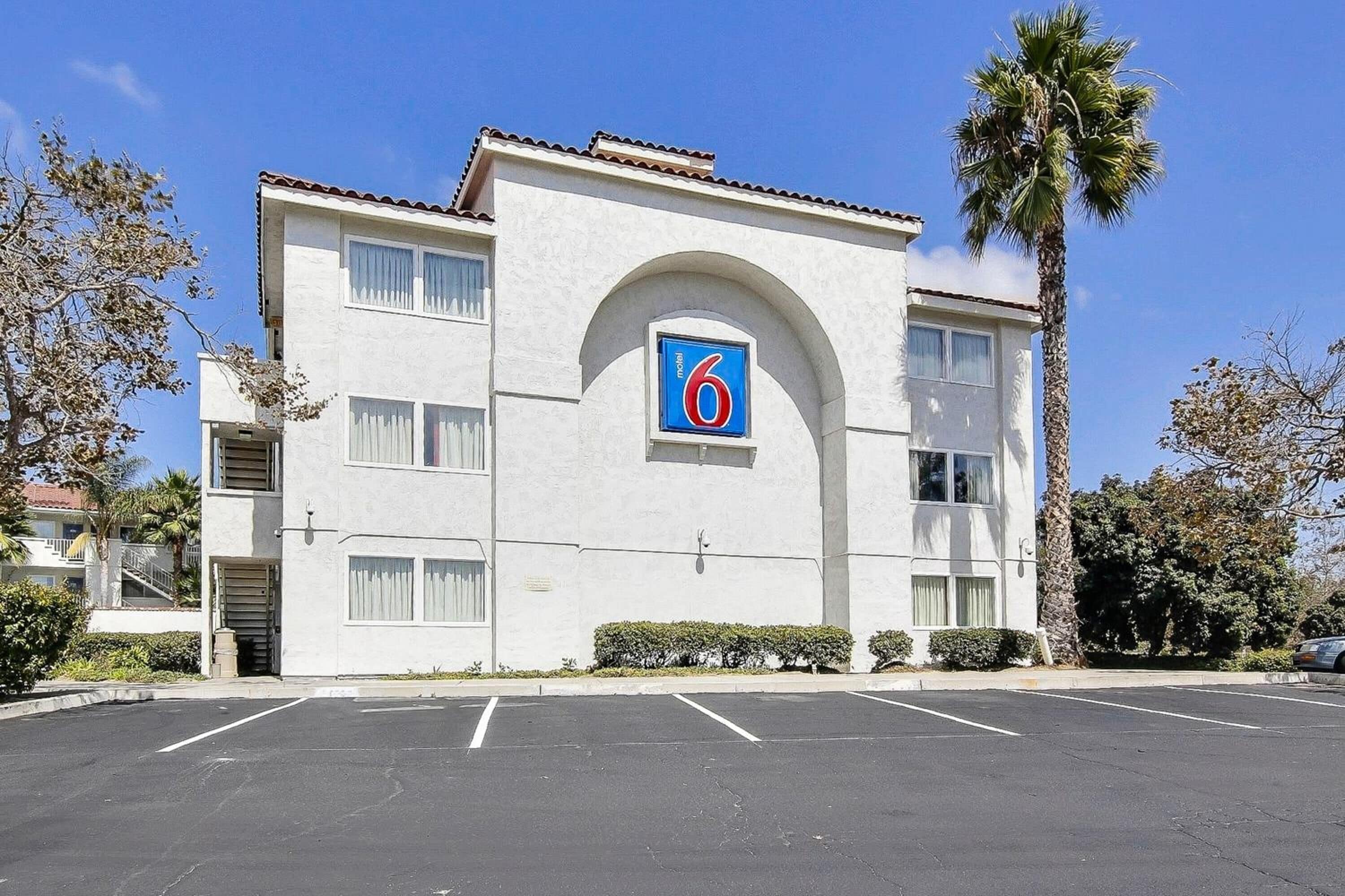 A 6-story motel with a central arched entrance, a parking lot in front, and palm trees around.