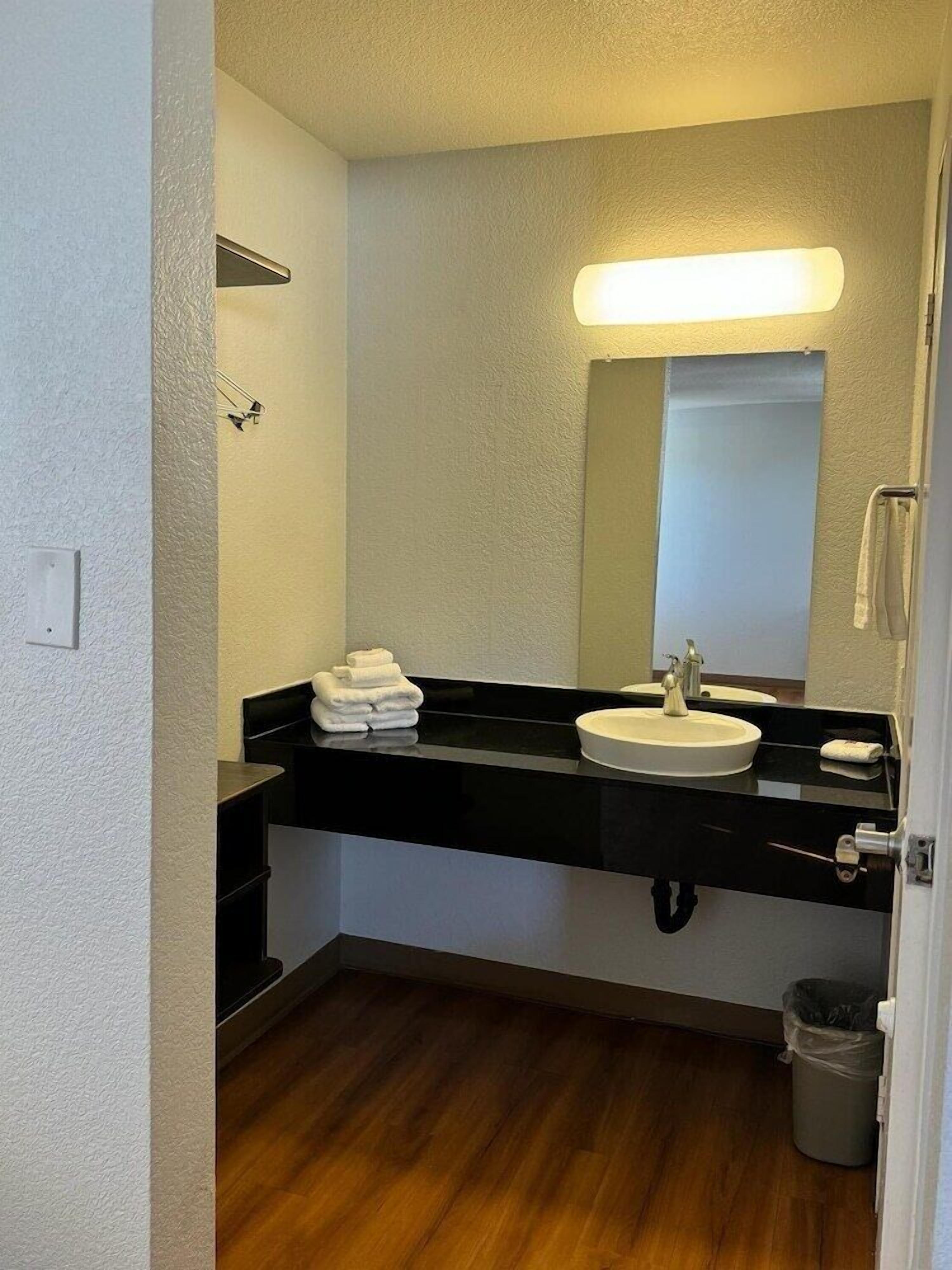 A bathroom with a black countertop, white sink, and a mirror. There are towels stacked on the countertop and a trash can in the corner.