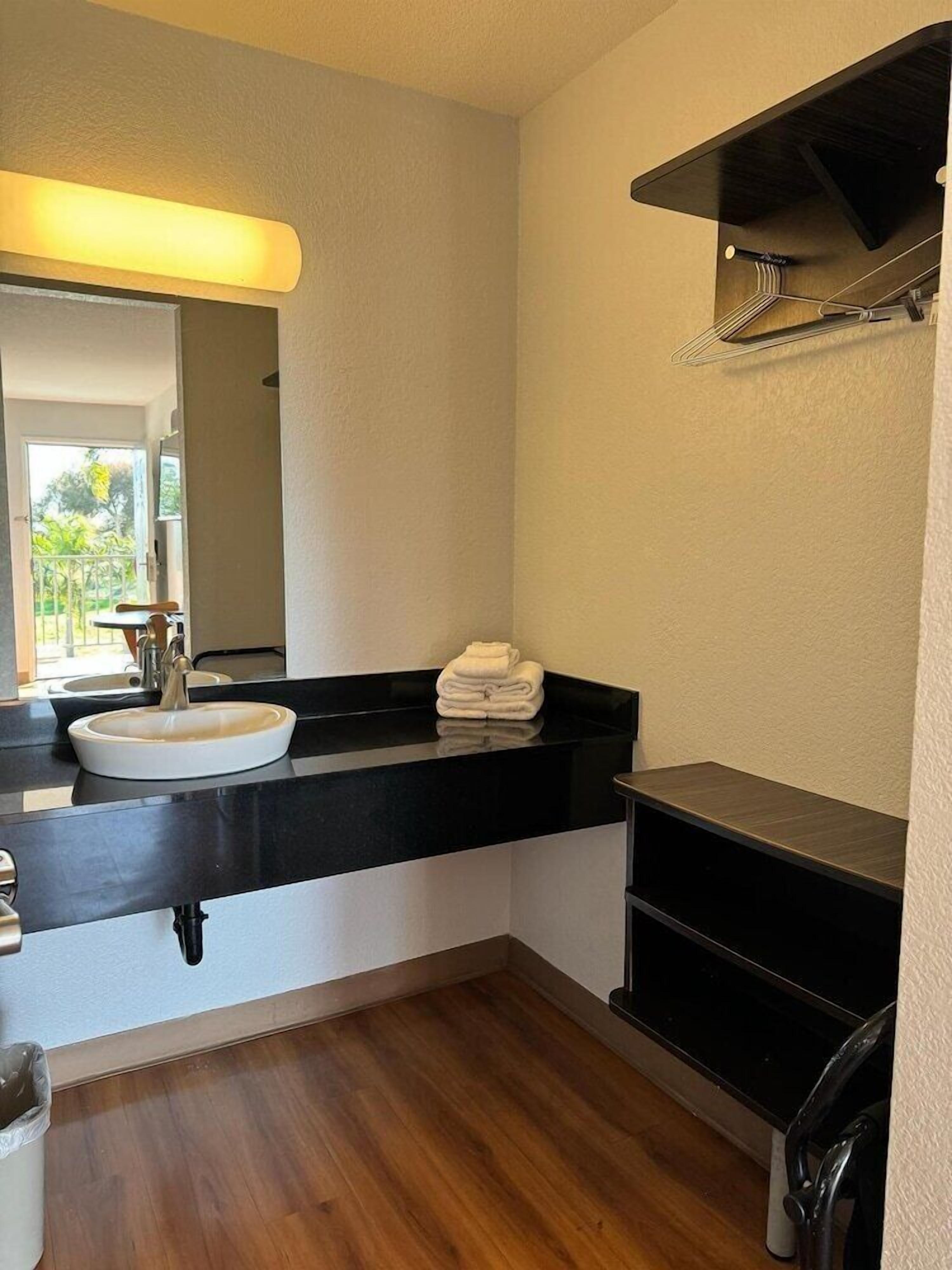 A bathroom with a white sink, black countertop, and a mirror. There is a towel stack on the countertop and a shelf above the sink.