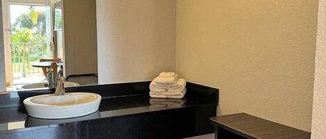Un baño con lavamanos blanco, encimera negra y un espejo. Hay una pila de toallas sobre la encimera y un estante encima del lavamanos.