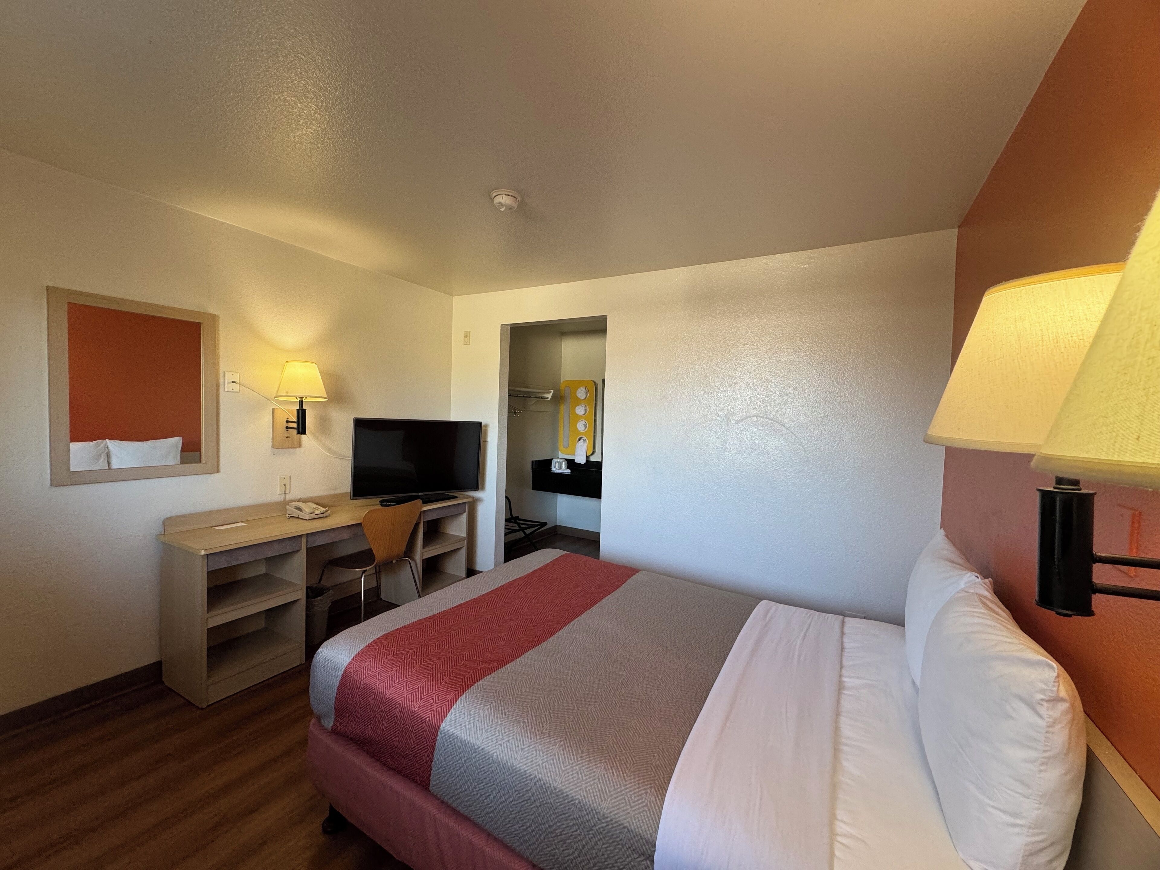 A hotel room with a bed, a desk with a TV, a mirror, and a lamp.