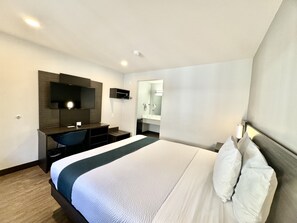 Habitación de hotel con cama, escritorio con silla, televisor de pantalla plana, espejo y baño visible a través de una puerta abierta.