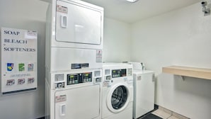 Een wasserette met gestapelde wasmachines en drogers, een plank met schoonmaakmiddelen, en een bord waarop beschikbare producten staan vermeld.