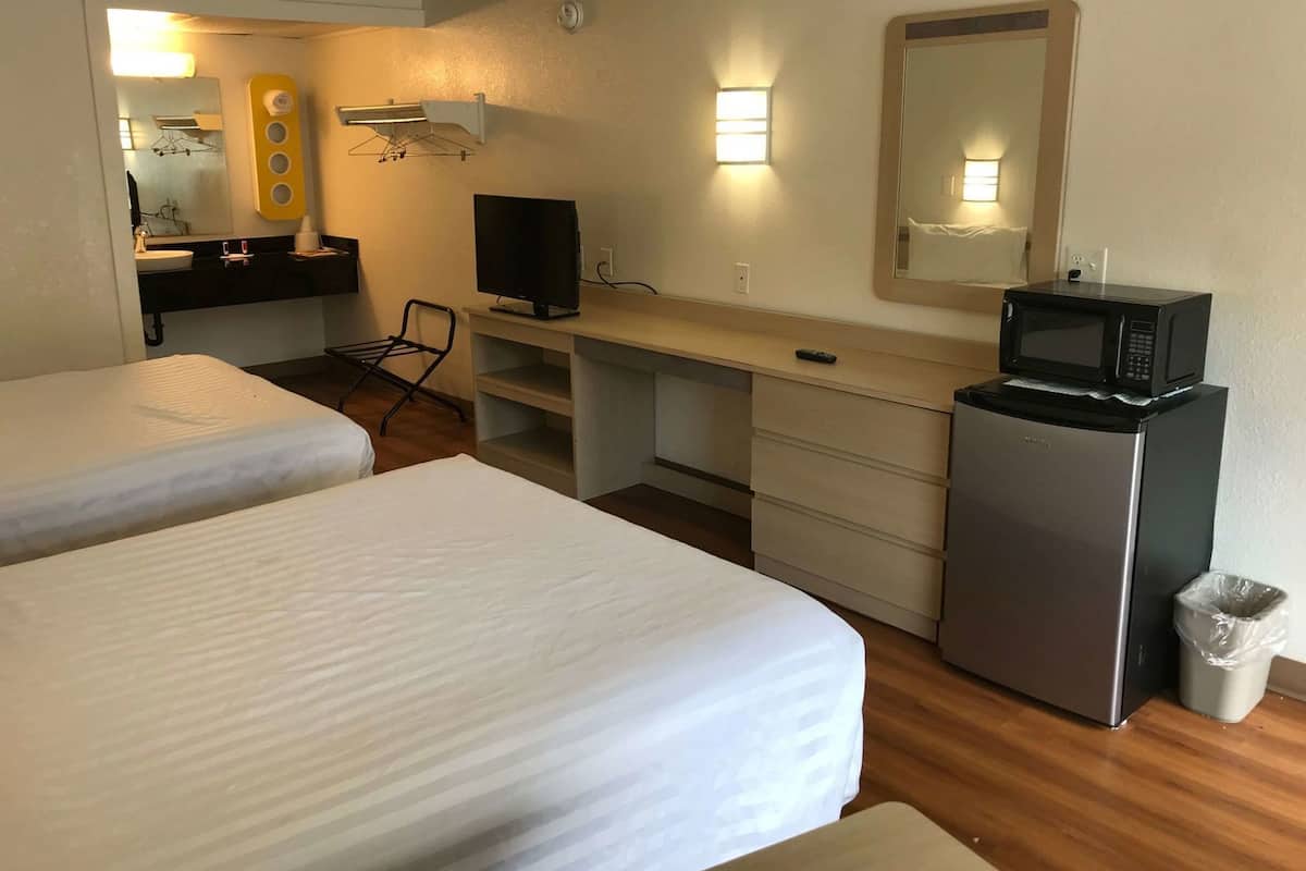 A hotel room with two beds, a desk with a TV, a microwave, a small trash bin, and a mirror on the wall.
