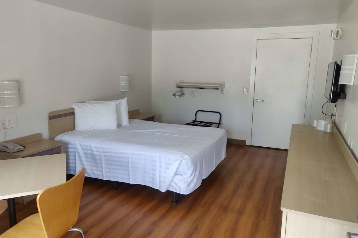 A hotel room with a bed, a desk, a chair, a bedside table, a wall-mounted TV, a radiator, and a door.