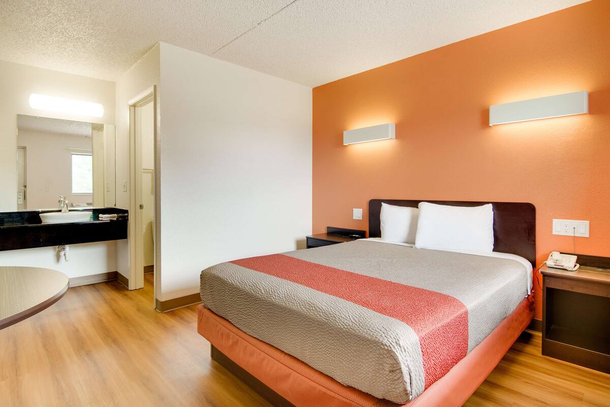 A hotel room with a bed, a bedside table, a nightstand, a mirror, a sink, and a wooden floor.
