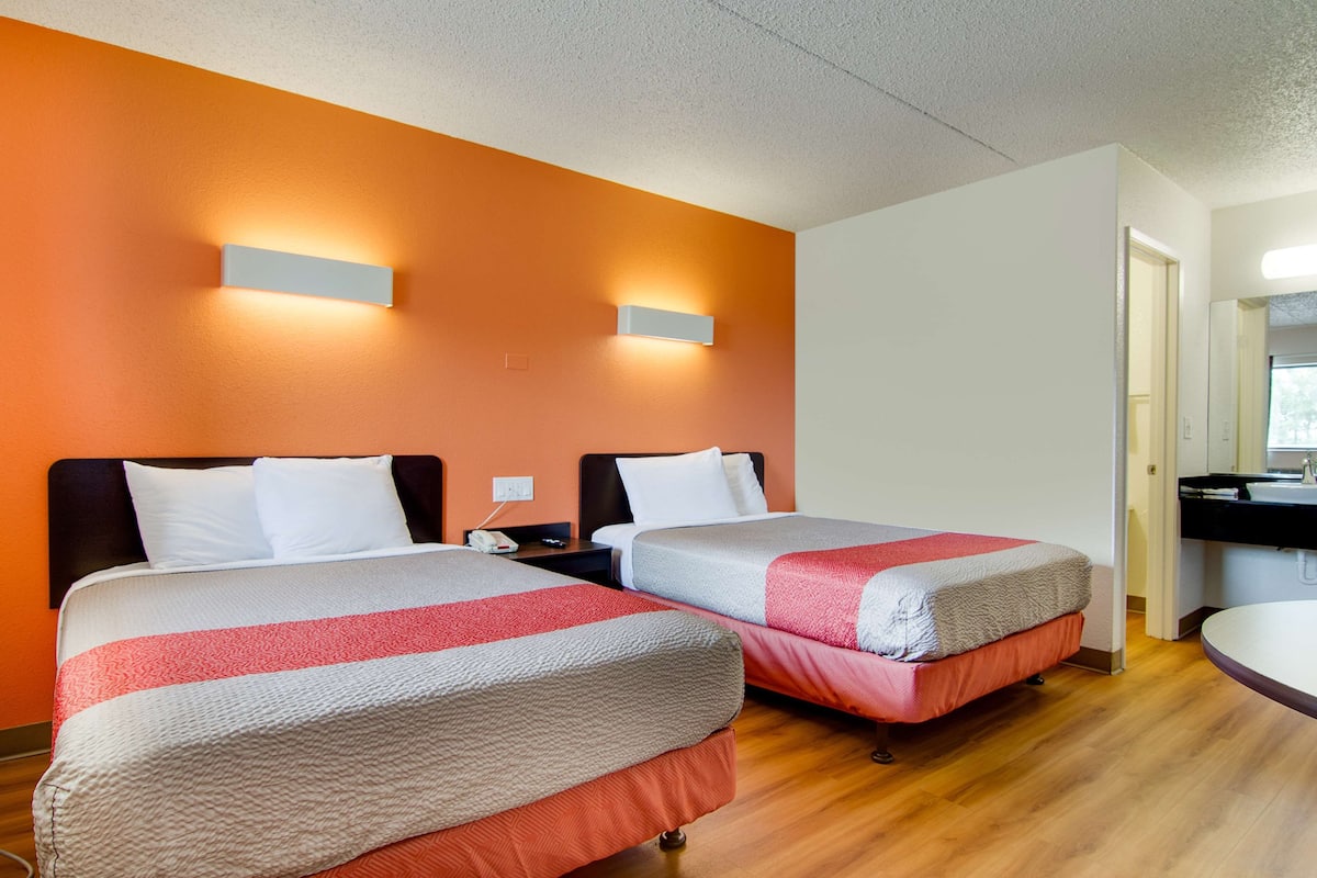 A hotel room with two beds, a wooden floor, a bathroom with a sink, and a mirror.