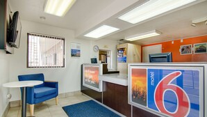 Vestíbulo de motel con mostrador de recepción, una silla azul, una mesita y un reloj en la pared.