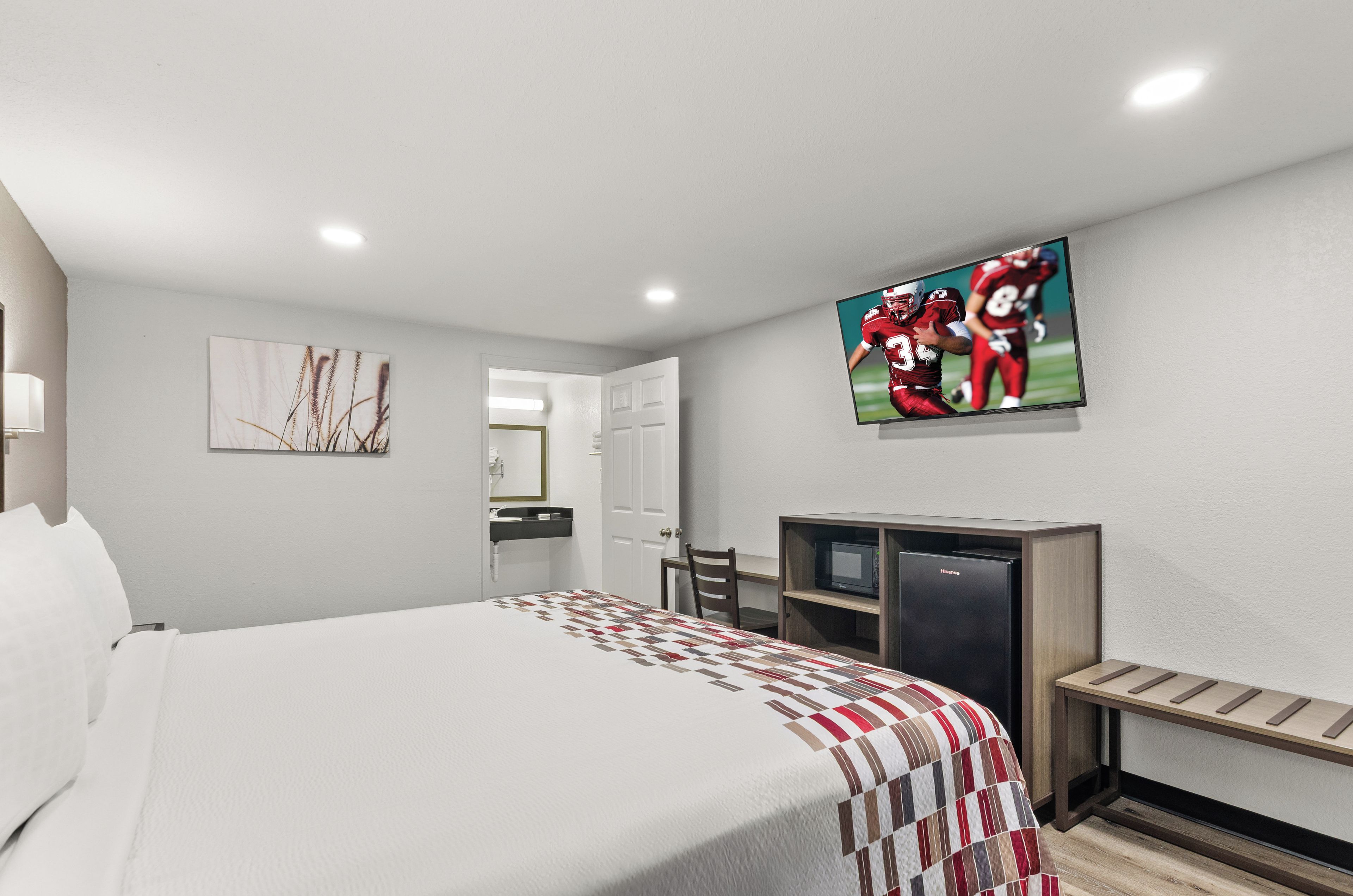 A hotel room with a bed, a television mounted on the wall, a desk with a microwave, and a door leading to a bathroom.