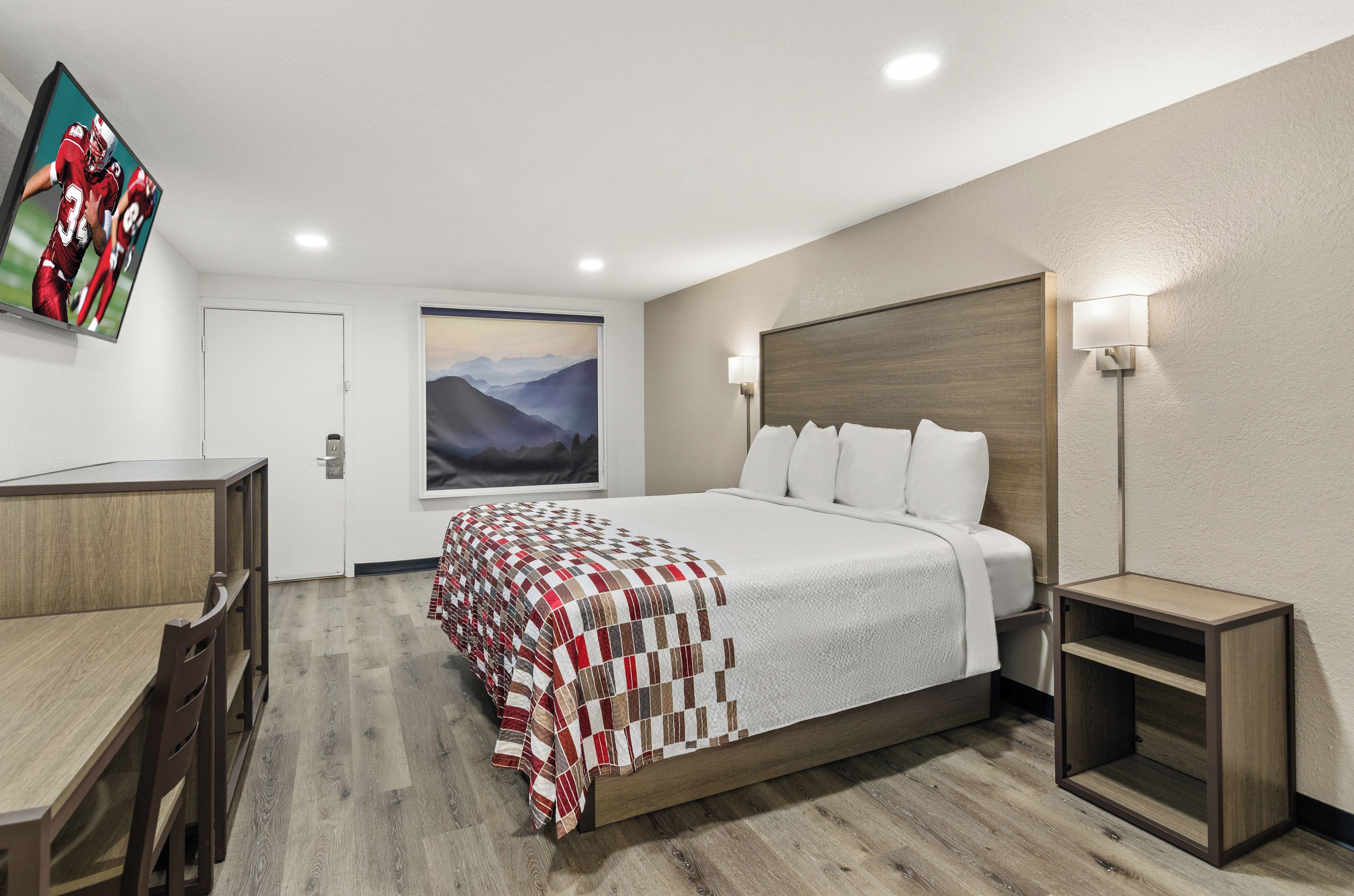 A hotel room with a bed, a desk, a chair, a painting, and a television on the wall.