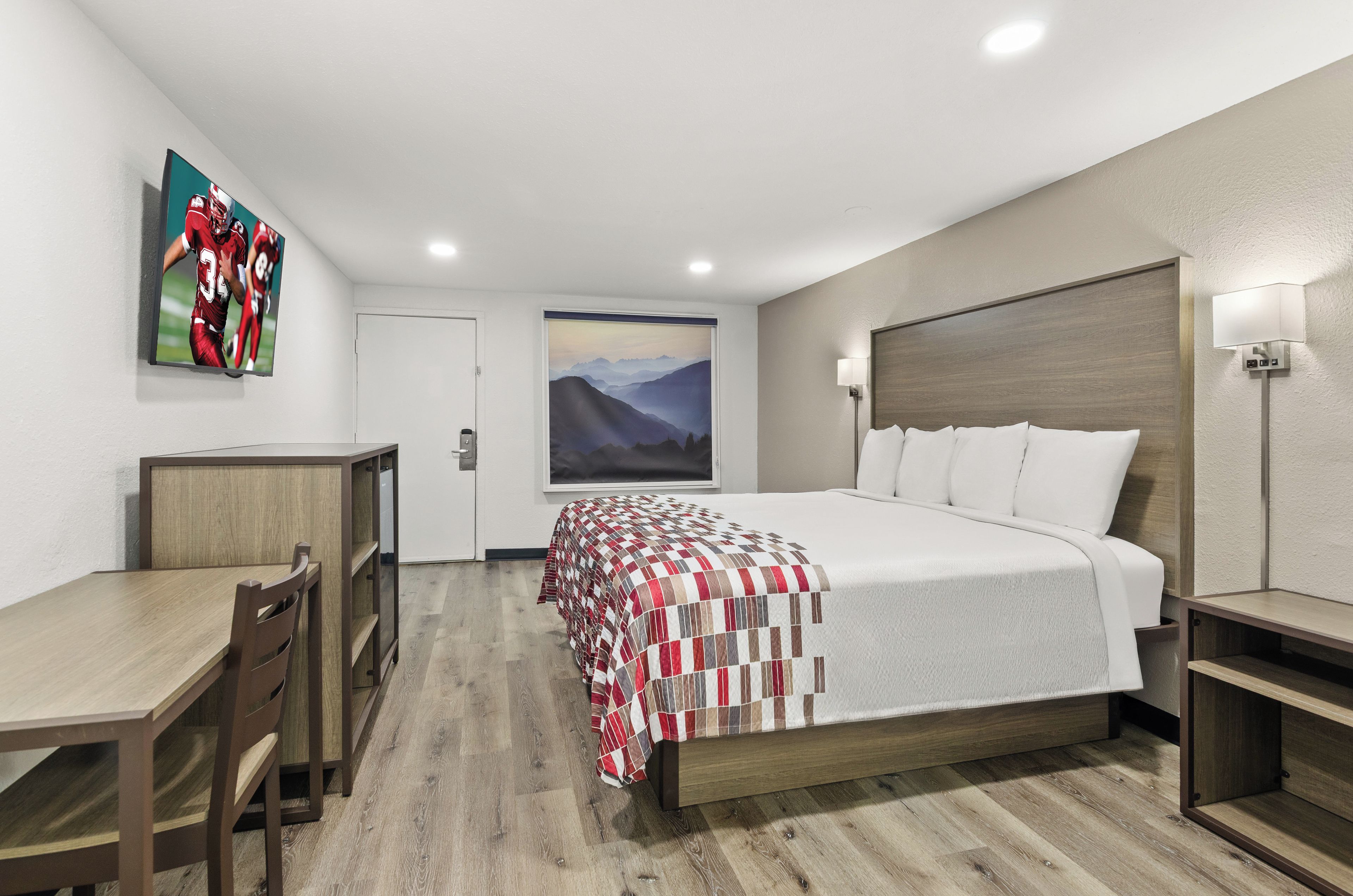 A hotel room with a bed, a desk, a chair, a painting on the wall, and a lamp.