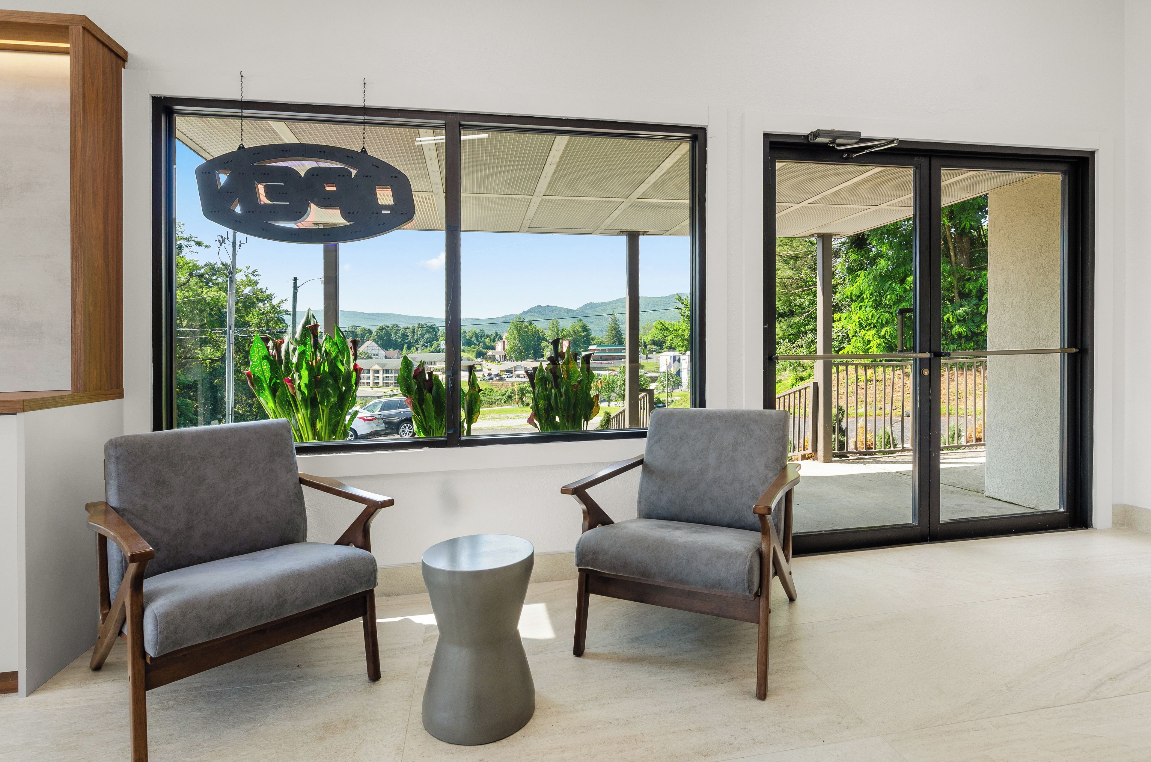 Ein modernes Interieur mit zwei grauen Sesseln, einem metallischen Couchtisch und großen Fenstern mit Blick auf Grünflächen und Gebäude.
