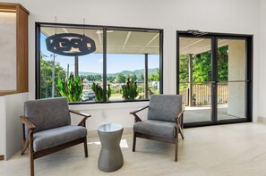 Un interior moderno con dos sillones grises, una mesa de centro metálica y ventanas amplias que ofrecen vistas a zonas verdes y edificios.