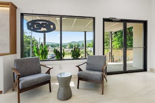 Um interior moderno com dois poltronas cinzas, uma mesa de centro metálica e grandes janelas com vista para vegetação e edifícios.