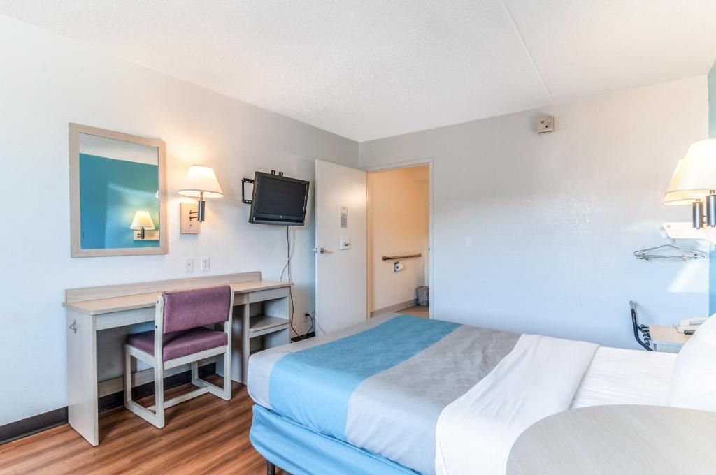 Habitación de hotel con cama, escritorio, silla, televisor y dos lámparas de pared.