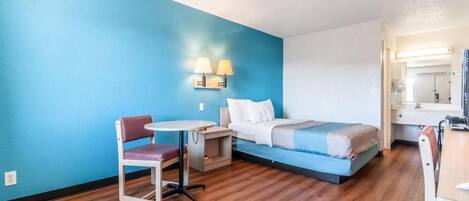 A hotel room with a bed, a desk, a chair, a small table, and a wall-mounted lamp.