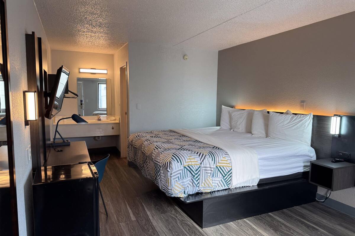 A modern hotel room with a large bed, bedside tables, a desk with a computer, a mirror, and a television.