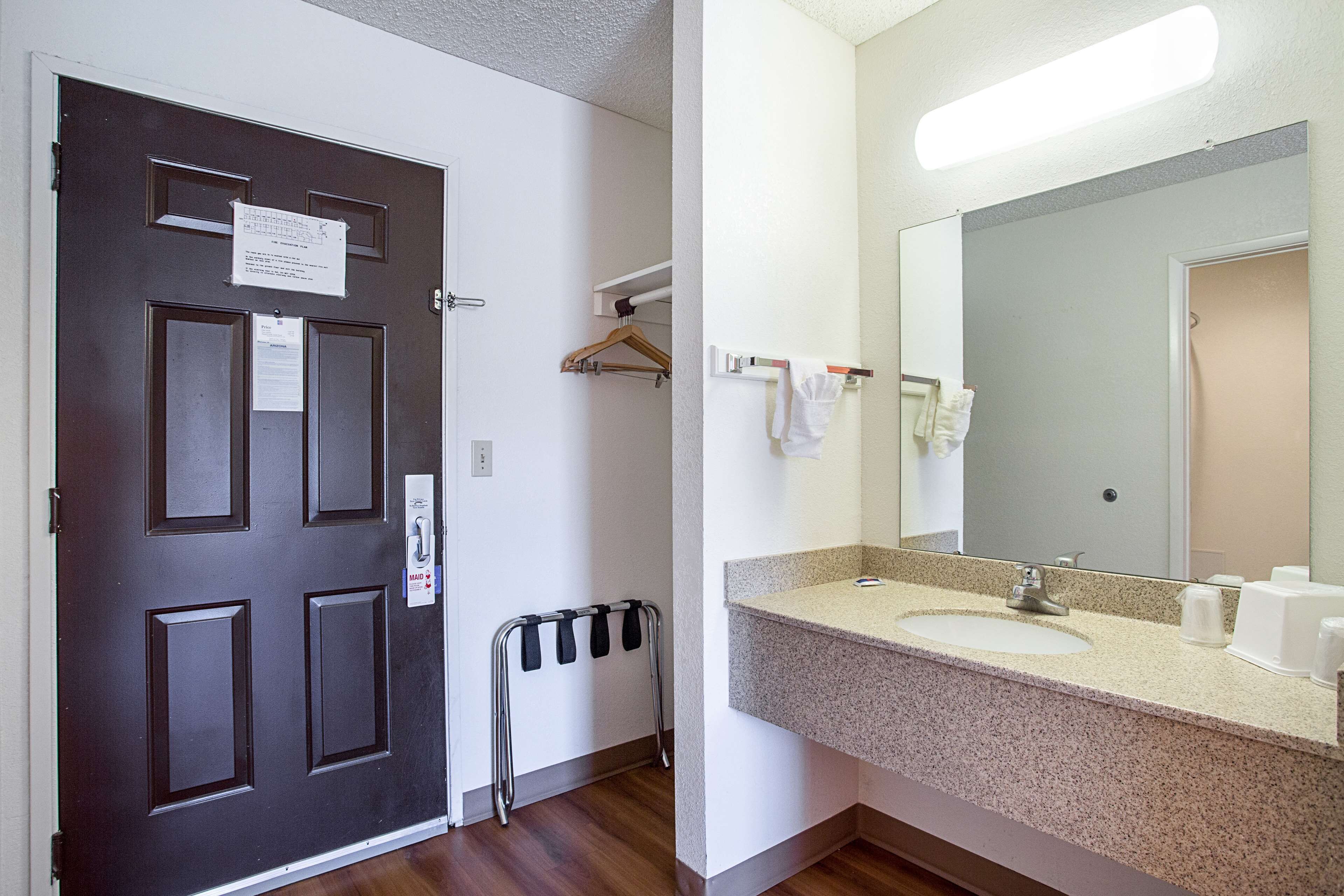 A hotel room with a dark wooden door, a white wall, a bathroom with a sink, mirror, and towel rack.