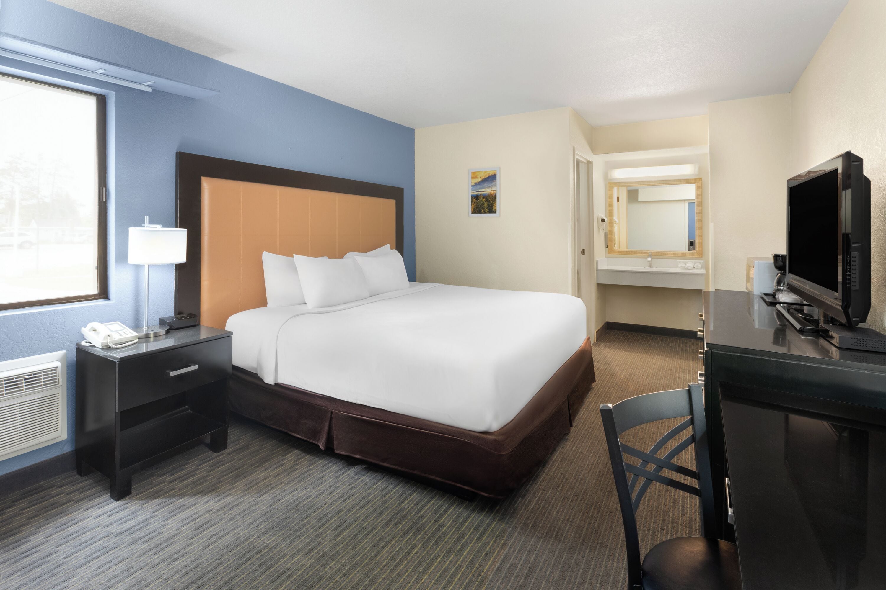 A hotel room with a bed, a desk with a computer, a chair, a nightstand with a phone, and a window.
