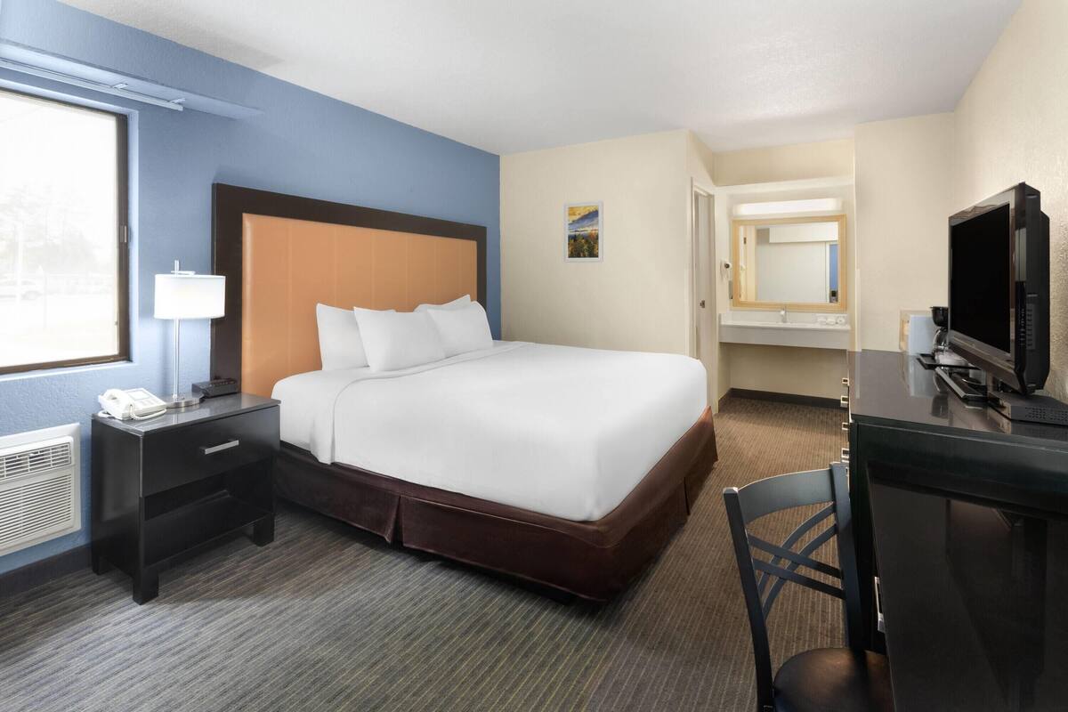Habitación de hotel con cama, escritorio con computadora, silla, mesita de noche con teléfono, lámpara y ventana.