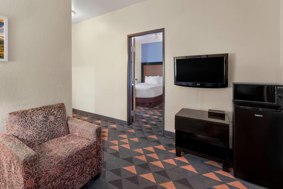 Habitación de hotel con sofá, televisor, refrigerador y cama visible a través de una puerta abierta.