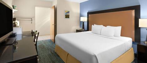 A hotel room with a large bed, a desk with a TV, a chair, a nightstand with a lamp, and a door leading to another room.