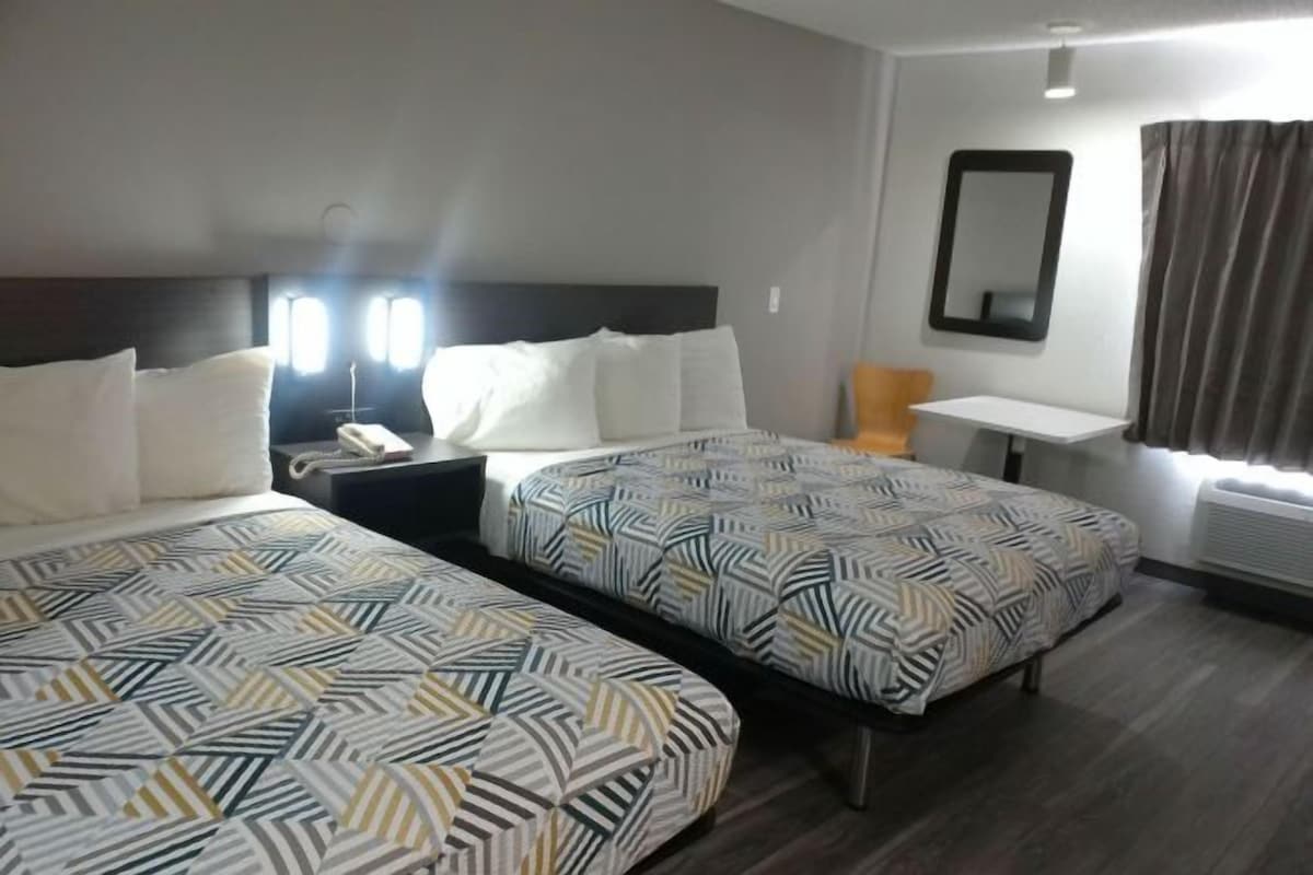A hotel room with two beds, a bedside table, a chair, a mirror, and a window with curtains.