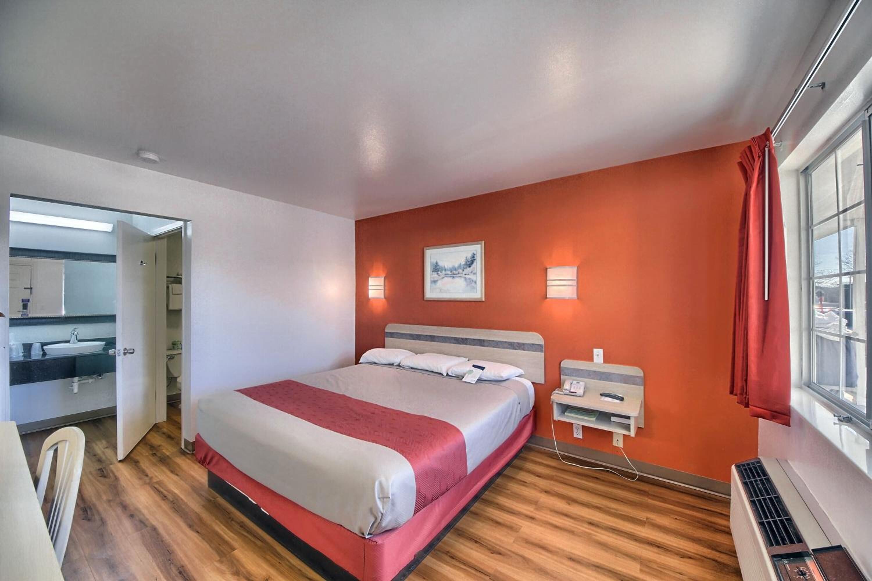A hotel room with a bed, a bedside table, a mirror, a window, and a wall-mounted air conditioner.