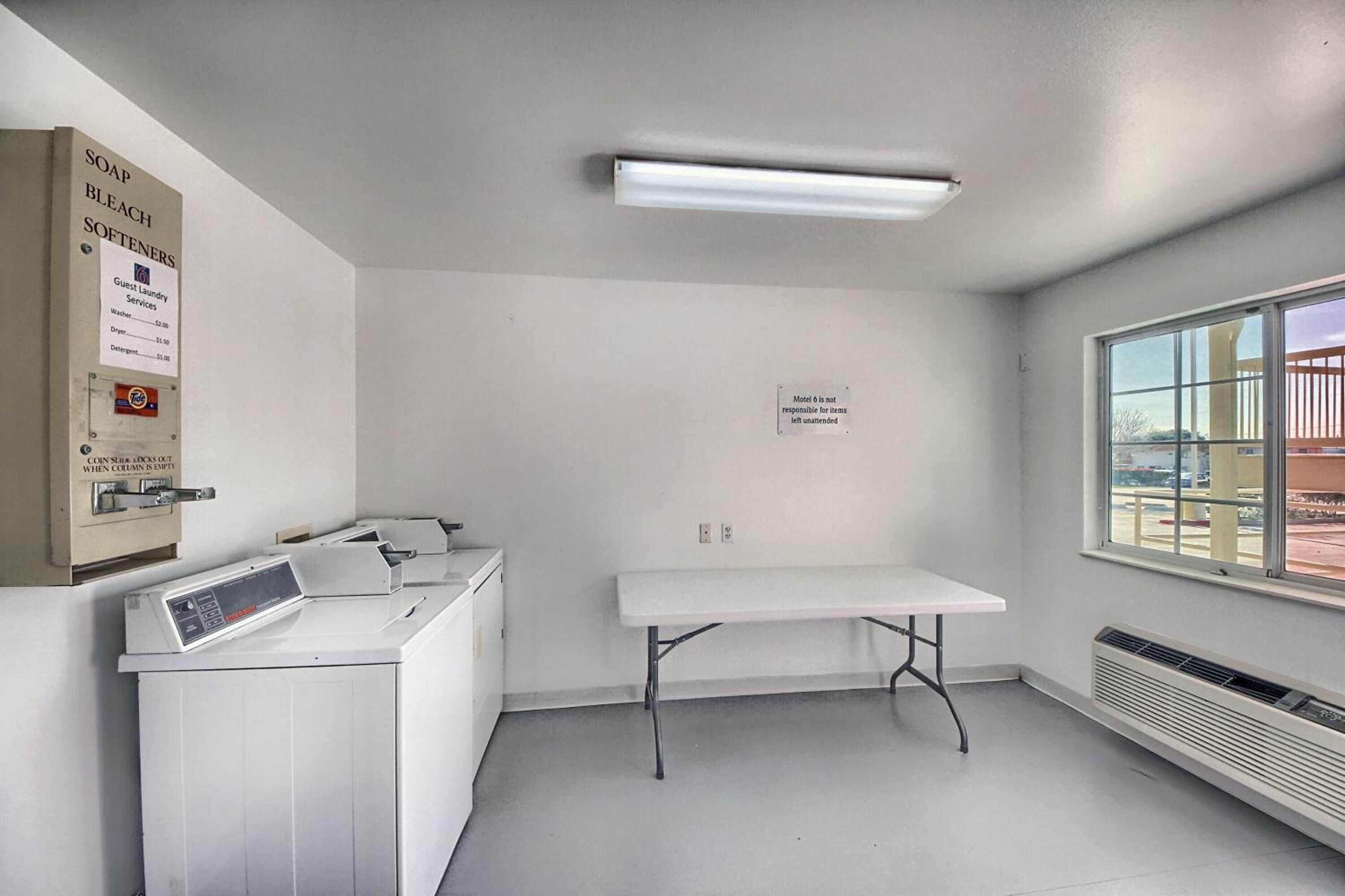 A hotel room with a washing machine, a table, a heater, and a window with a view of the outside.