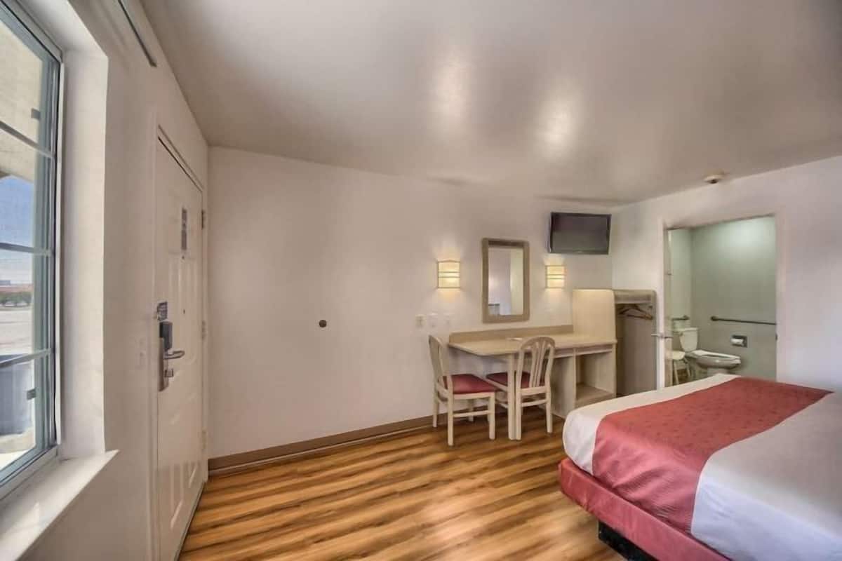 A hotel room with a bed, a desk with a chair, a TV mounted on the wall, and a window.