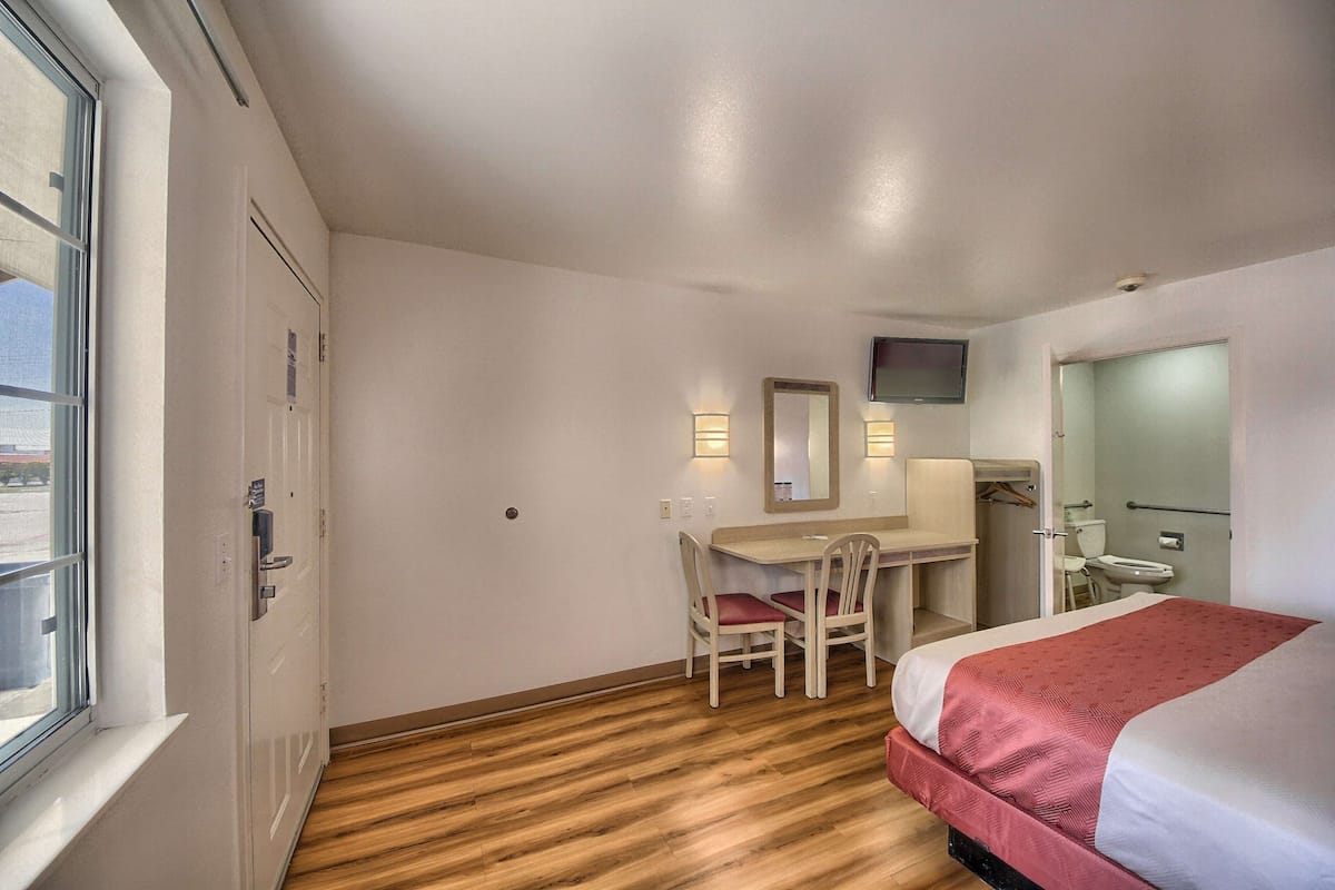 A hotel room with a bed, a desk with a chair, a television mounted on the wall, and a window.