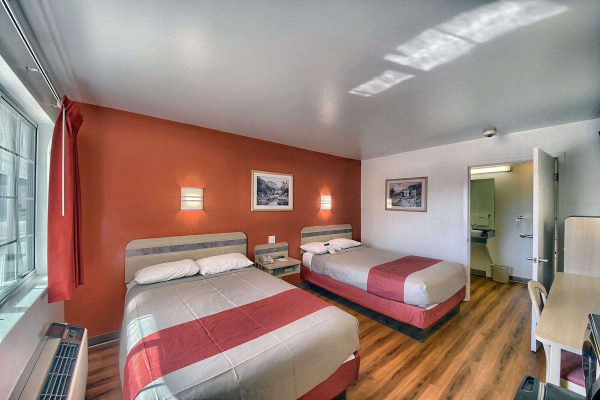 A hotel room with two beds, a desk, and a chair. There are two wall-mounted lights and a window with curtains.