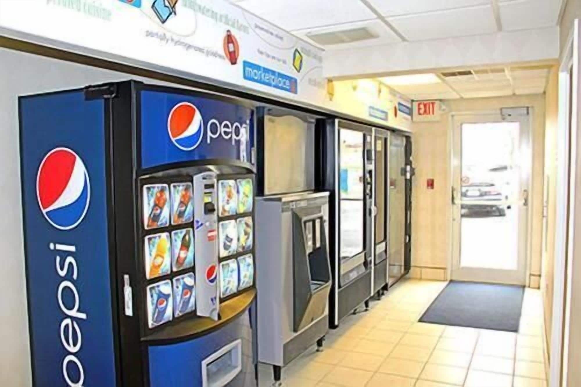 A row of Pepsi vending machines in a hallway with a tiled floor and an exit door visible in the background.