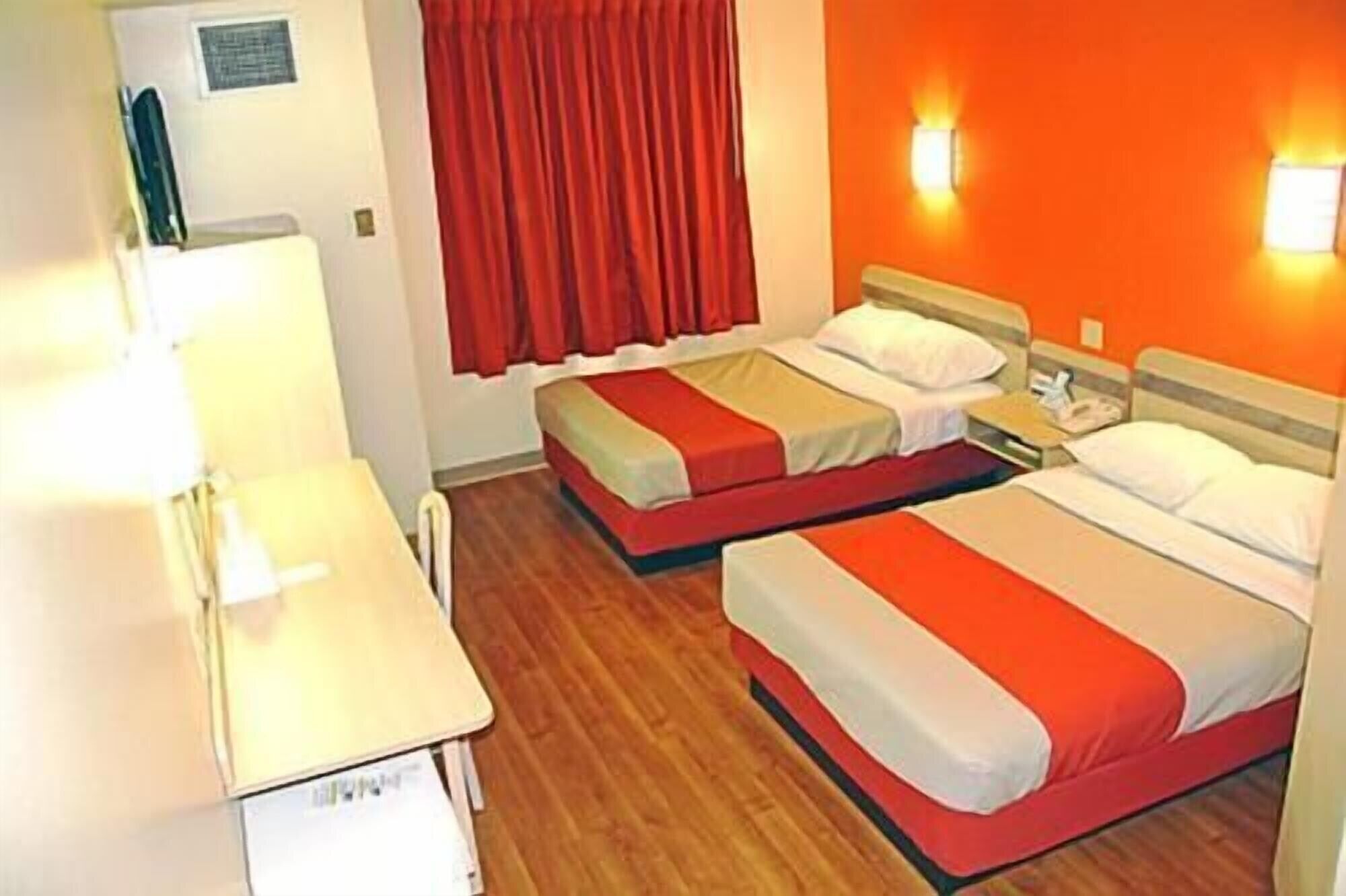 A hotel room with two beds, a desk, and a window with red curtains.