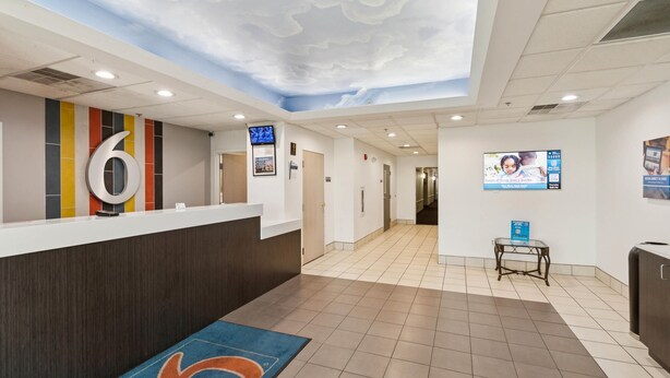 A hotel reception area with a modern design, including a reception desk, a large number '6' sign, a digital screen, and a blue carpet with a logo. - Motel 6 Milan, OH - Sandusky (Milan)