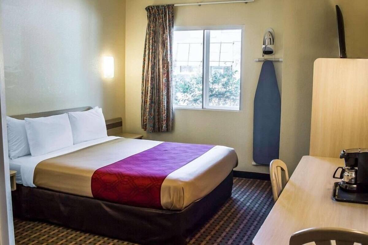 A hotel room with a bed, a table, a chair, a window with curtains, and a wall-mounted ironing board.