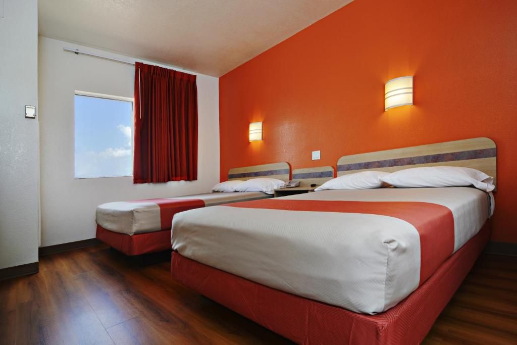 Quarto de hotel com duas camas, janela com cortinas vermelhas e luminárias fixadas na parede.