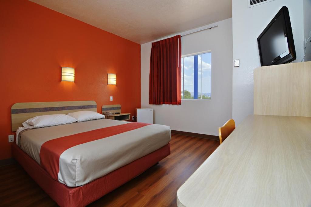 Quarto de hotel com cama, escrivaninha, televisão e janela com cortinas.