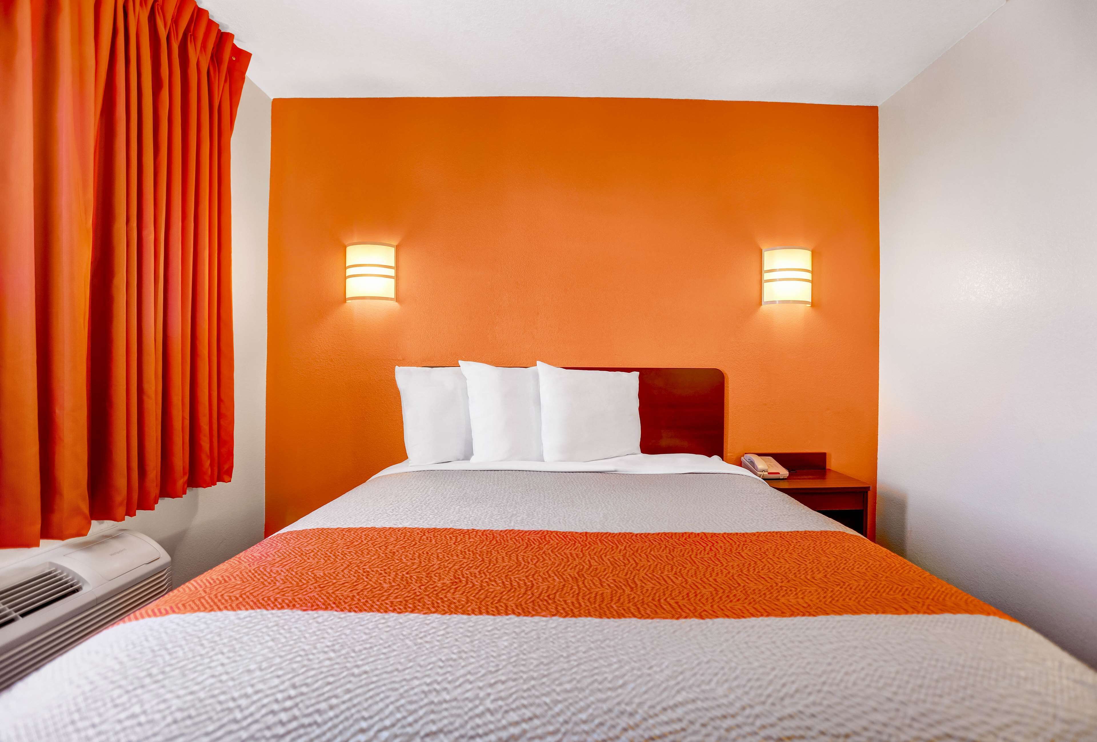 A hotel room with an orange accent wall, a bed with white and orange bedding, two wall-mounted lights, and a telephone on a nightstand.