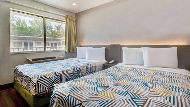 A hotel room with two beds, a window with curtains, a bedside table, and a wall-mounted air conditioning unit. - Motel 6 Port Allen, LA - Baton Rouge (Port Allen)