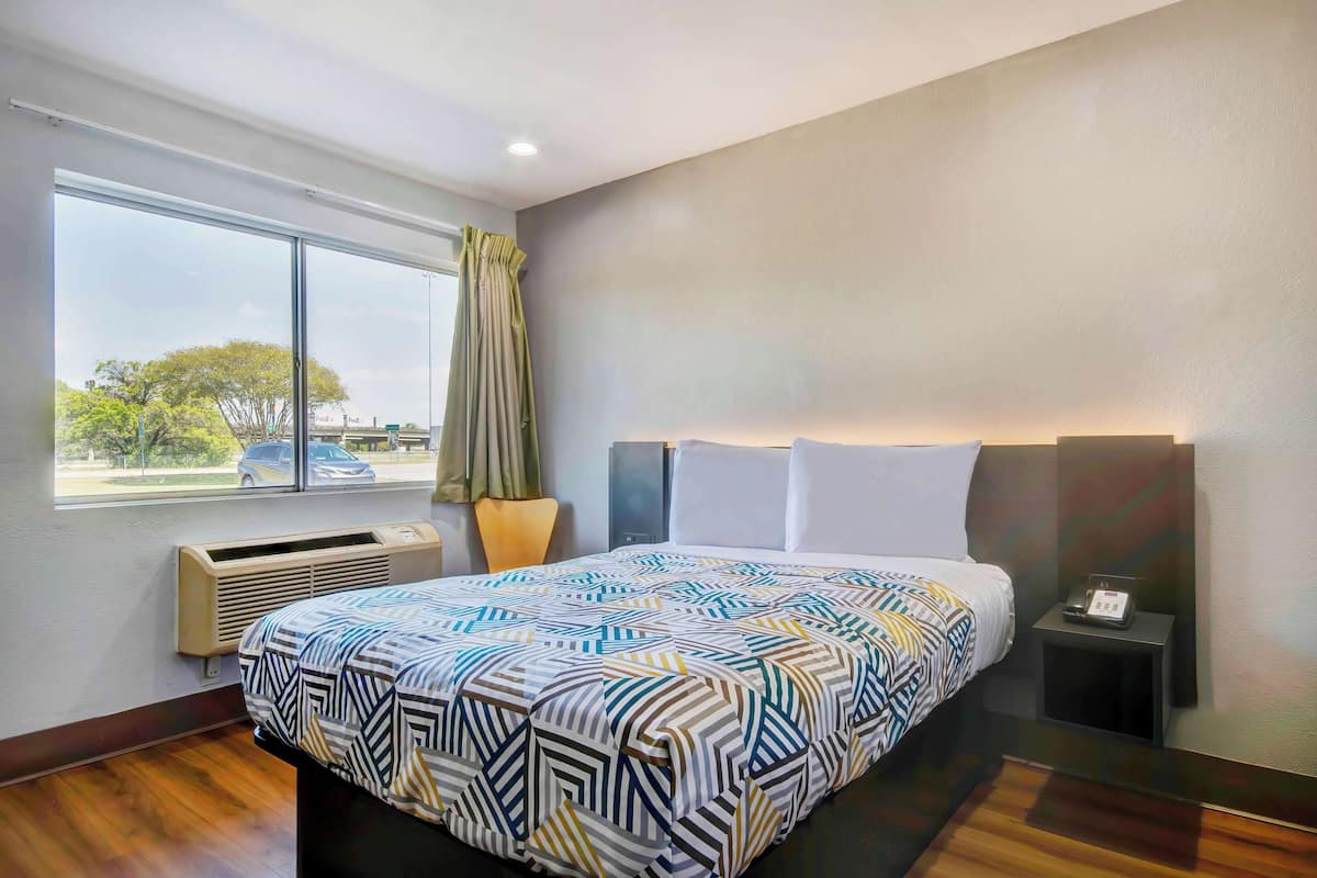 Ein Hotelzimmer mit einem großen Bett, einem Fenster mit Vorhängen, einem Nachttisch mit Telefon und einer wandmontierten Klimaanlage.