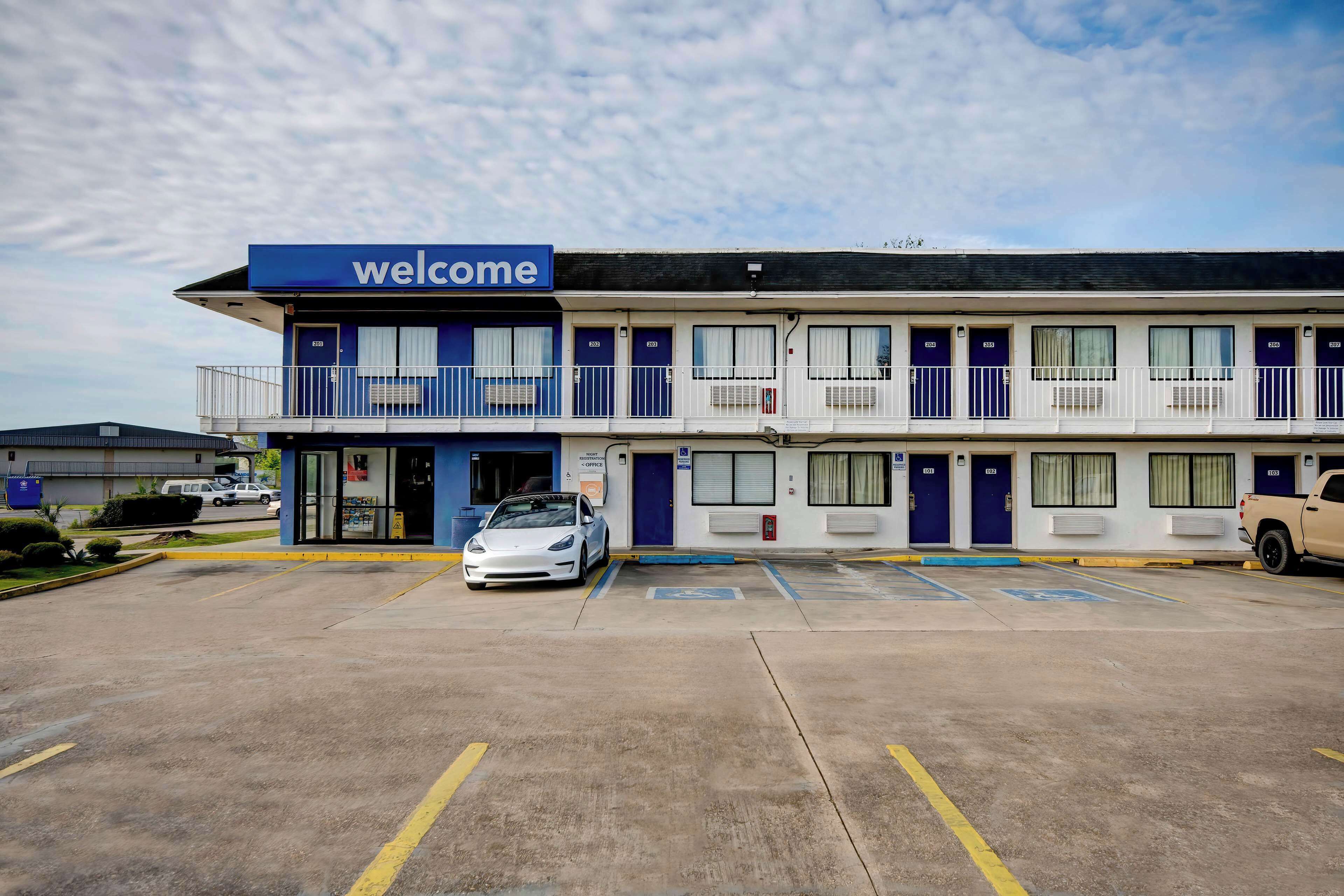 A motel with a 'welcome' sign, multiple rooms, a parking lot, and a white car parked in front.