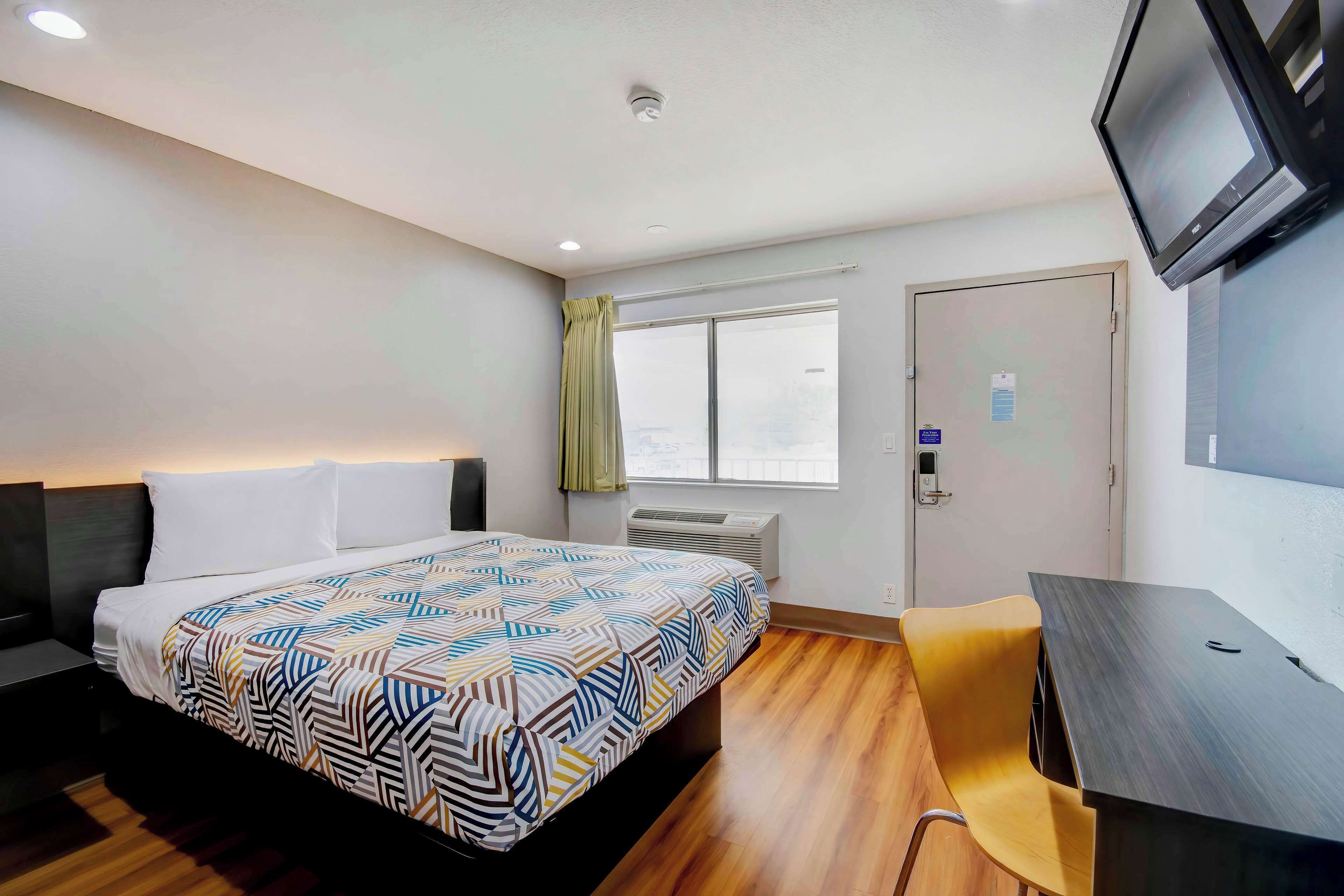A hotel room with a bed, a desk, a chair, a television, a window, and an air conditioning unit.