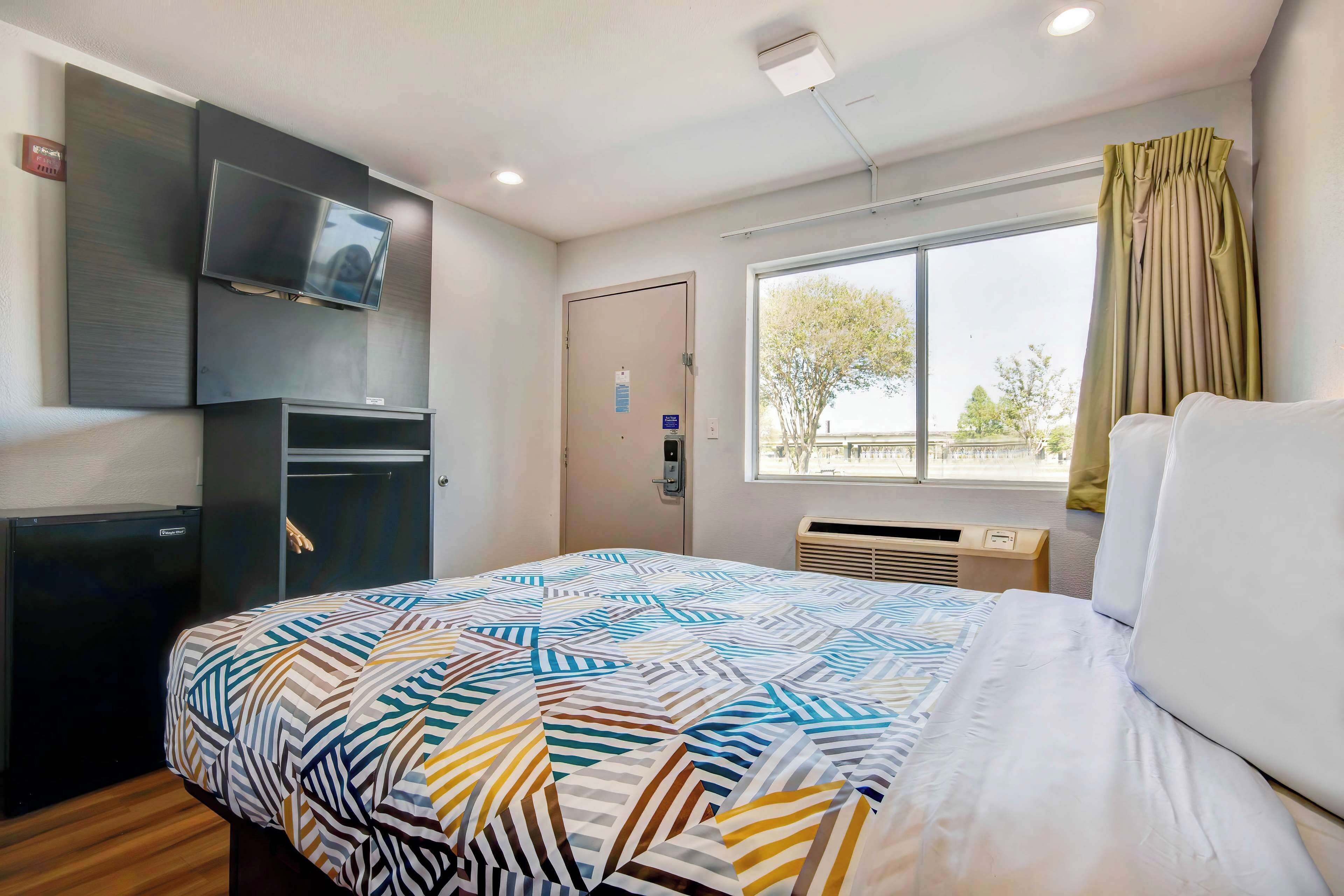 Habitación de hotel con cama, televisión montada en la pared, un refrigerador pequeño y una ventana con cortinas.