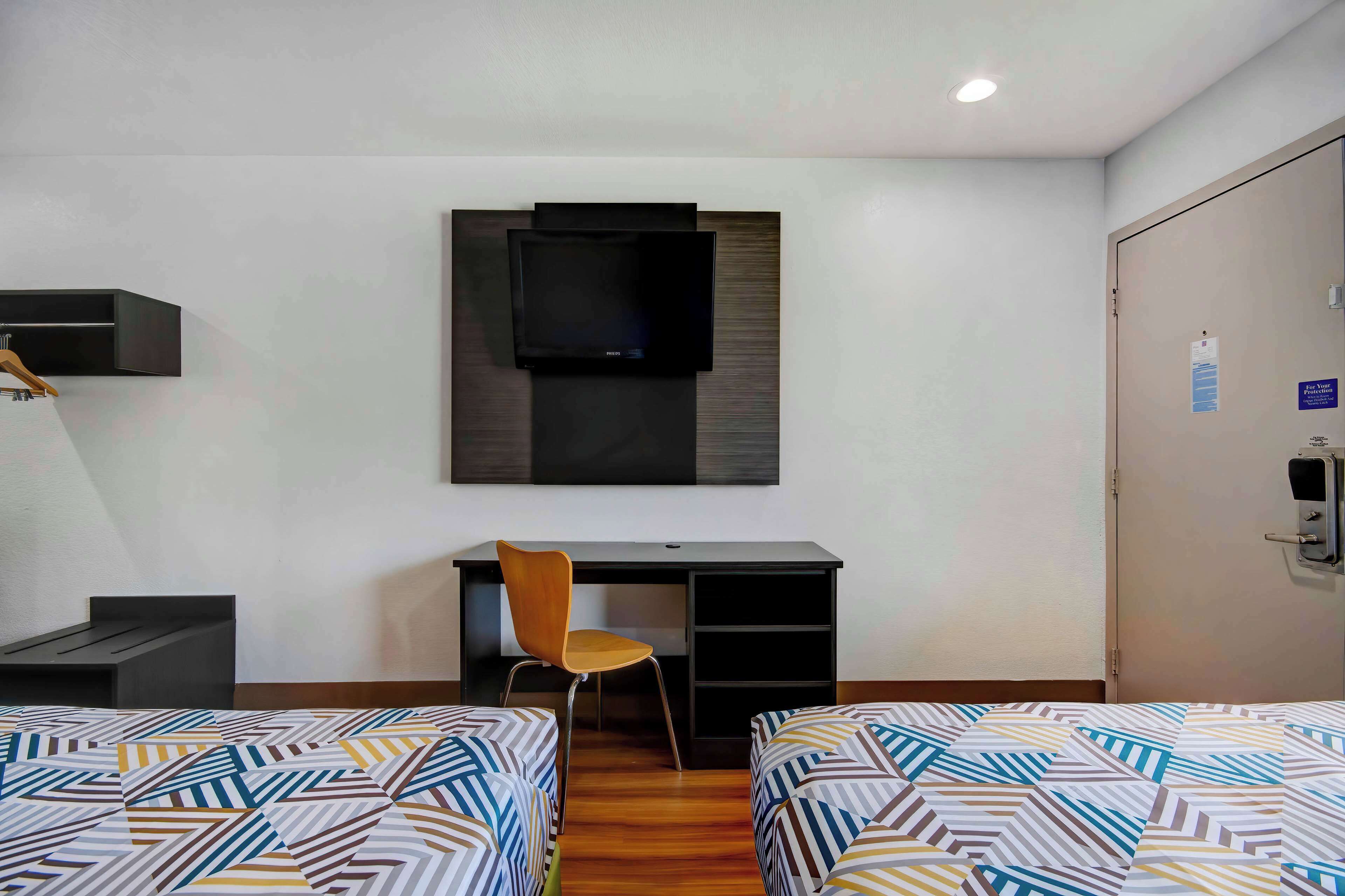 A hotel room with two beds, a desk, a chair, a TV mounted on the wall, and a door with a sign.