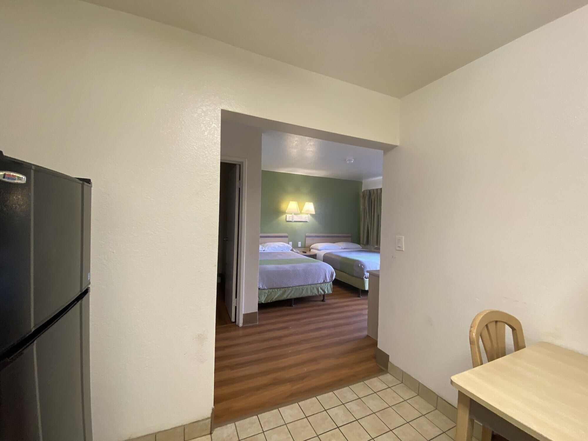 A compact hotel room with a kitchenette, two beds, a small dining table, and a wooden floor.
