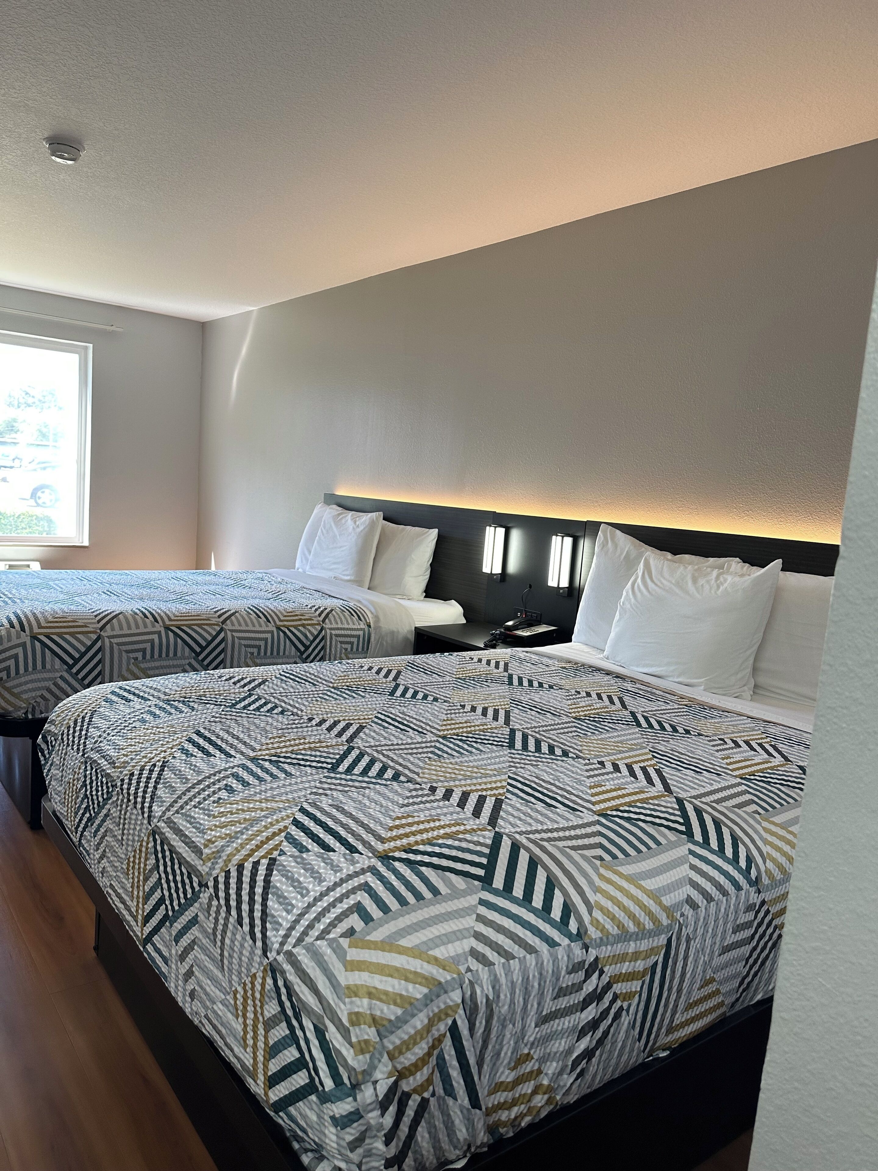 A hotel room with two beds, a large window, and a headboard with built-in lighting.