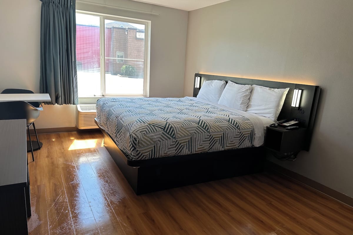 A modern hotel room with a wooden floor, a bed with patterned bedding, a desk with a chair, and a window with curtains.