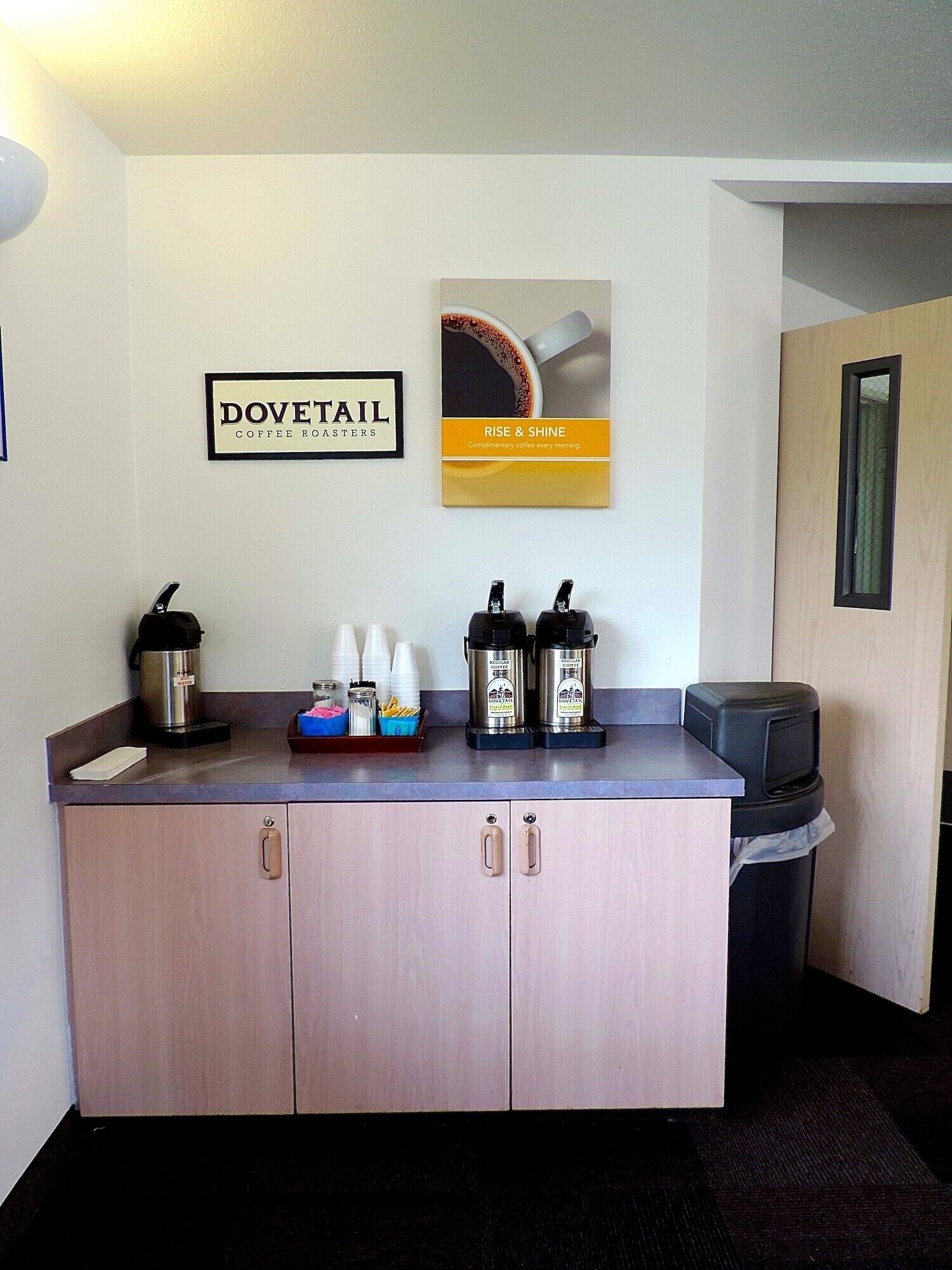 Eine Küchenzeile mit Kaffeemaschine, zwei Kaffeeautomaten und einem Papierkorb. An der Wand befinden sich zwei gerahmte Schilder, eines für 'Dovetail Coffee Roasters' und das andere für 'Rise & Shine'.