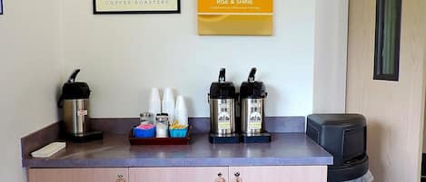 Eine Küchenzeile mit Kaffeemaschine, zwei Kaffeeautomaten und einem Papierkorb. An der Wand befinden sich zwei gerahmte Schilder, eines für 'Dovetail Coffee Roasters' und das andere für 'Rise & Shine'.